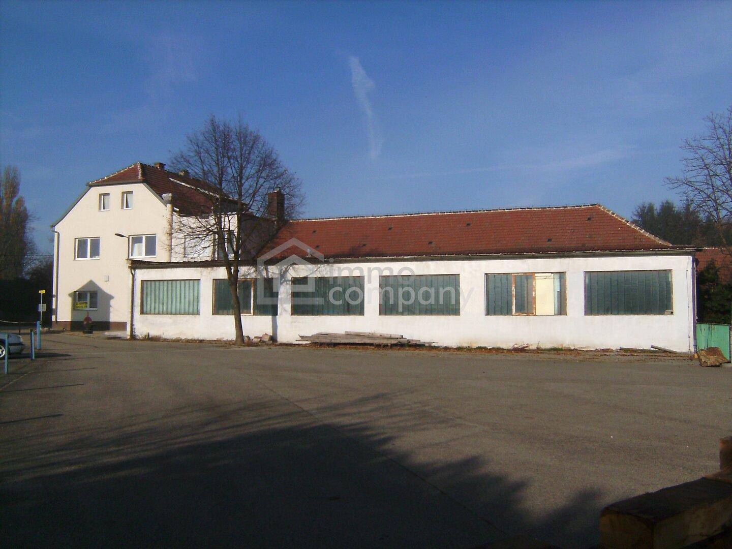 Weißes Gebäude mit rotem Ziegeldach und großen Fenstern, davor eine weitläufige asphaltierte Fläche.