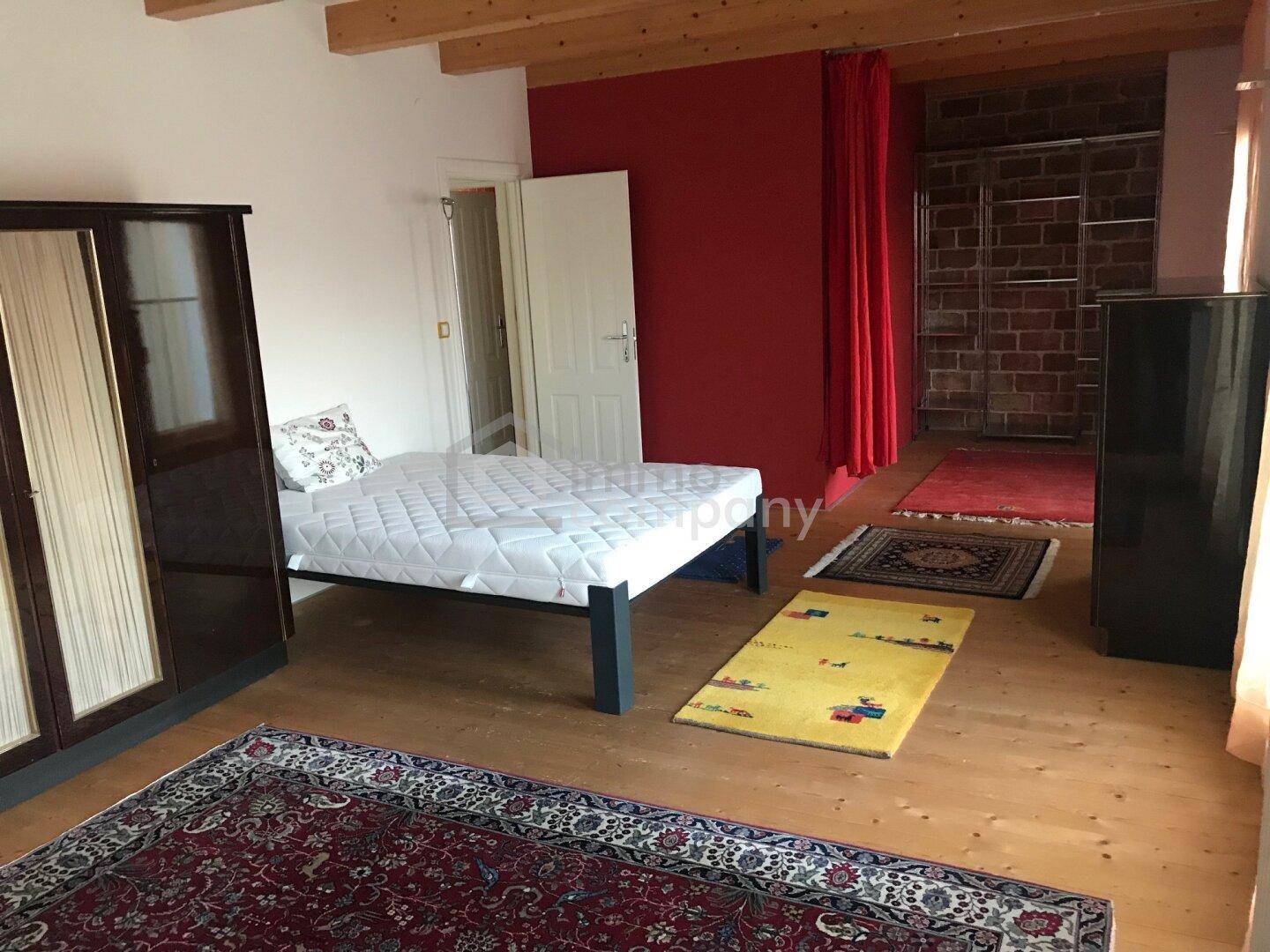 Schlafzimmer mit Holzboden, freiliegenden Holzbalken und einem roten Vorhang, der einen Bereich abtrennt.