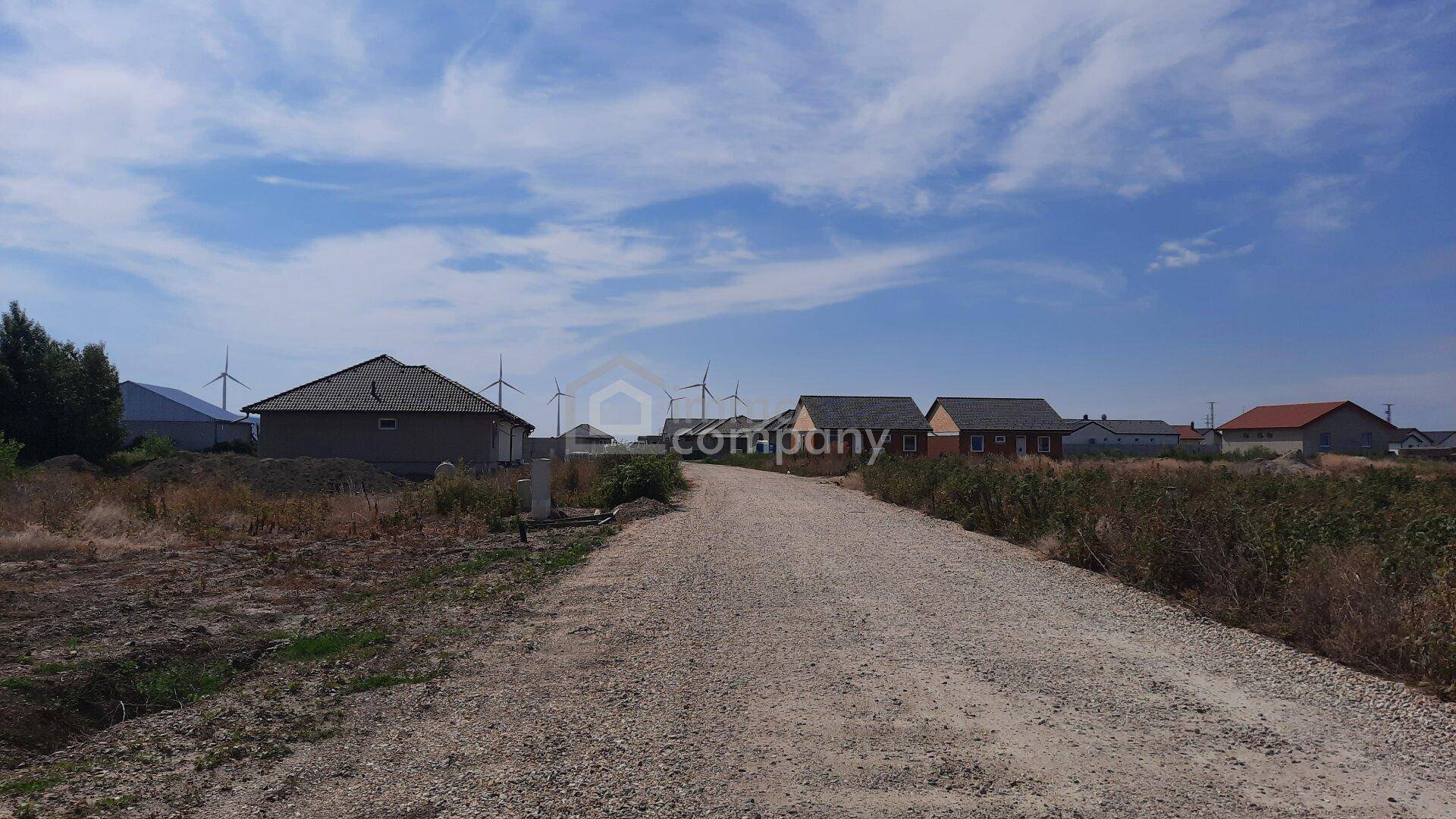 Schotterweg führt durch ländliche Landschaft mit Häusern und Windrädern am Horizont unter blauem Himmel.