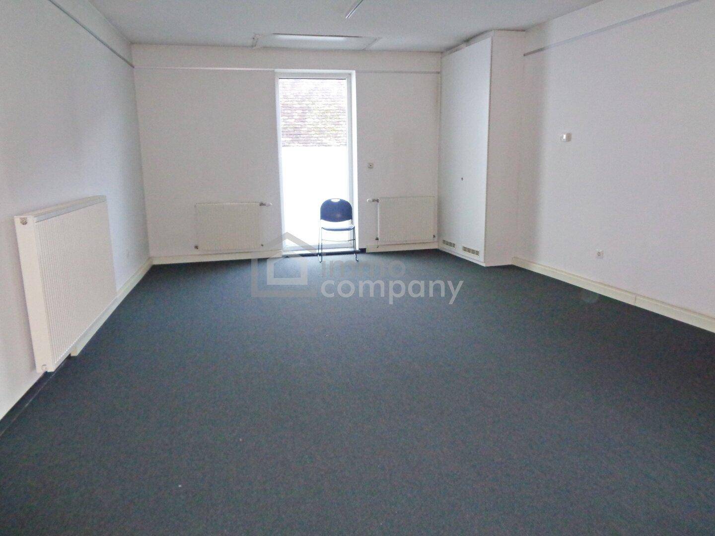 Leerer Büroraum mit Teppichboden, weißen Wänden und einem Fenster mit Heizkörpern.