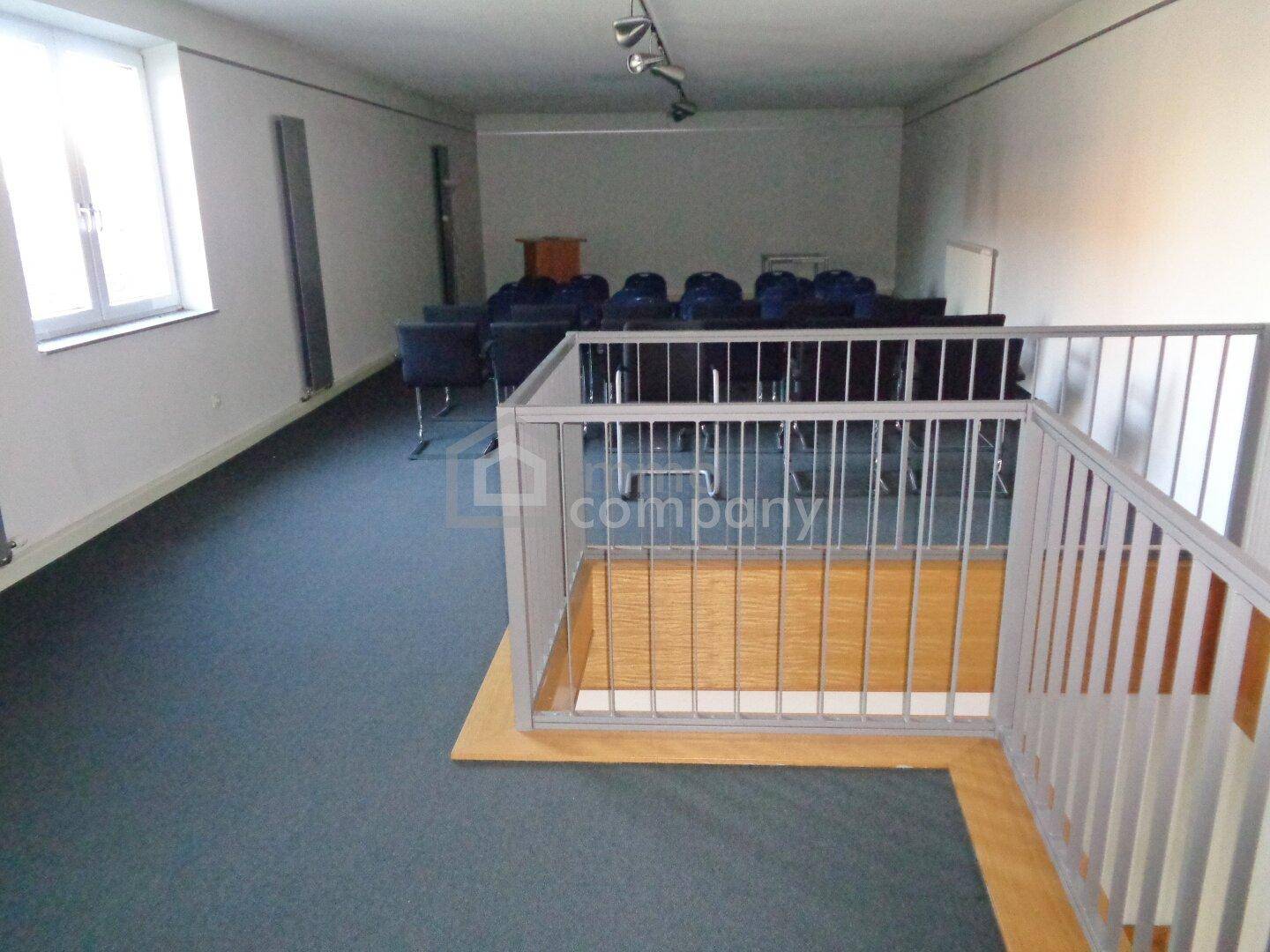 Großer Konferenzraum mit Teppichboden, Stühlen und einem Geländer, das einen tieferliegenden Bereich abgrenzt.