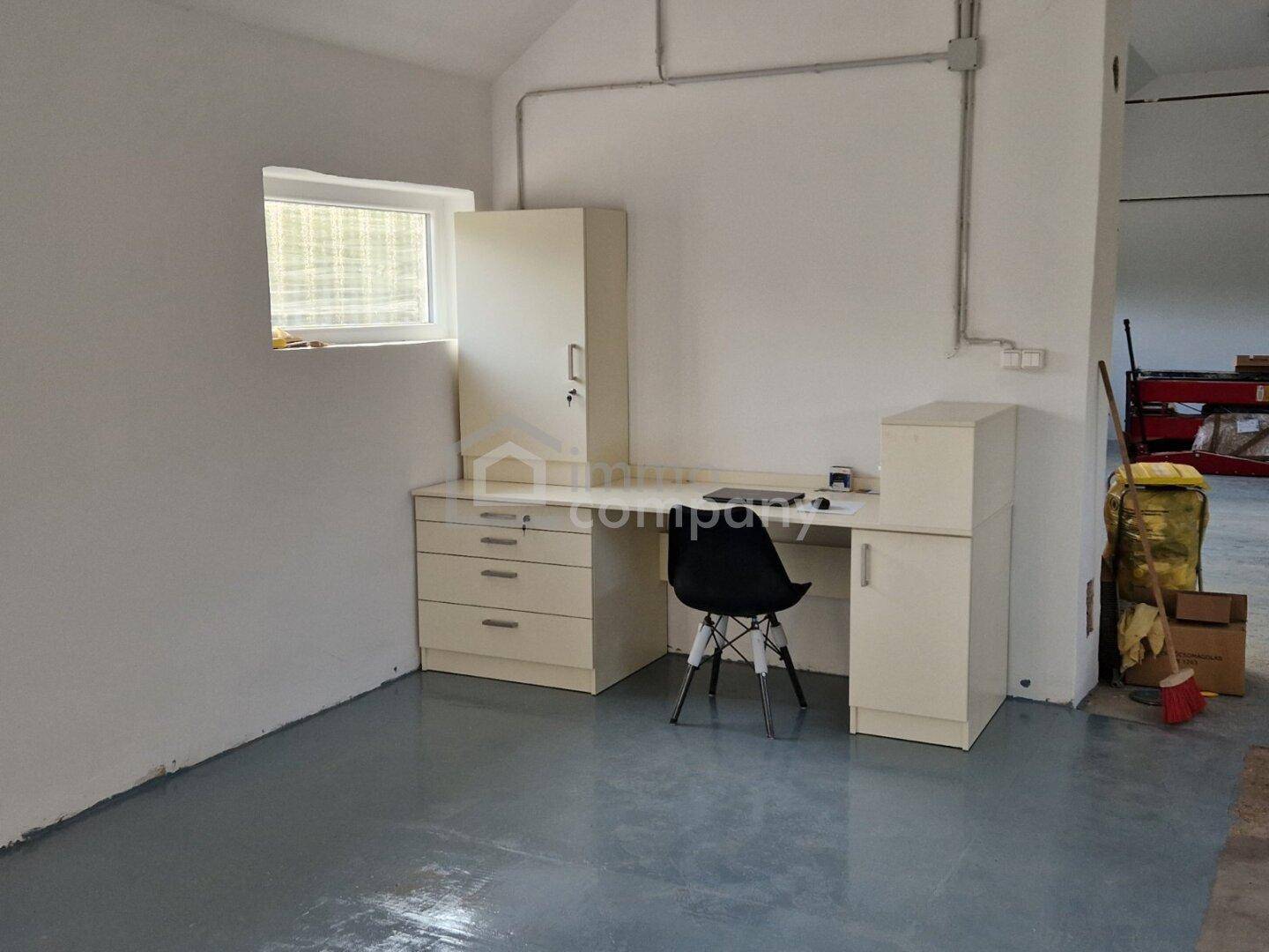 Kleiner Bürobereich mit Schreibtisch und Schränken in einem hellen Raum mit Fenster und grauem Boden.