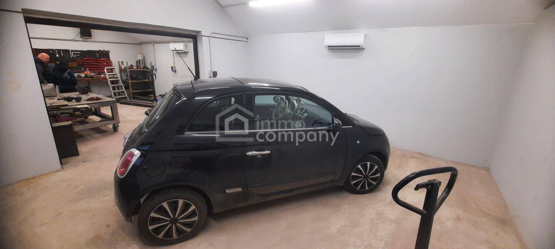 Garagenbereich mit einem schwarzen Kleinwagen, einem Klimagerät an der Wand und Zugang zu weiteren Räumen.