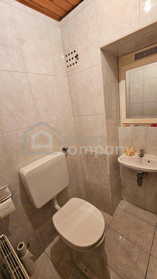 Kleines WC mit hellbeigen Fliesen, Toilette, kleinem Waschbecken und Holzdecke.