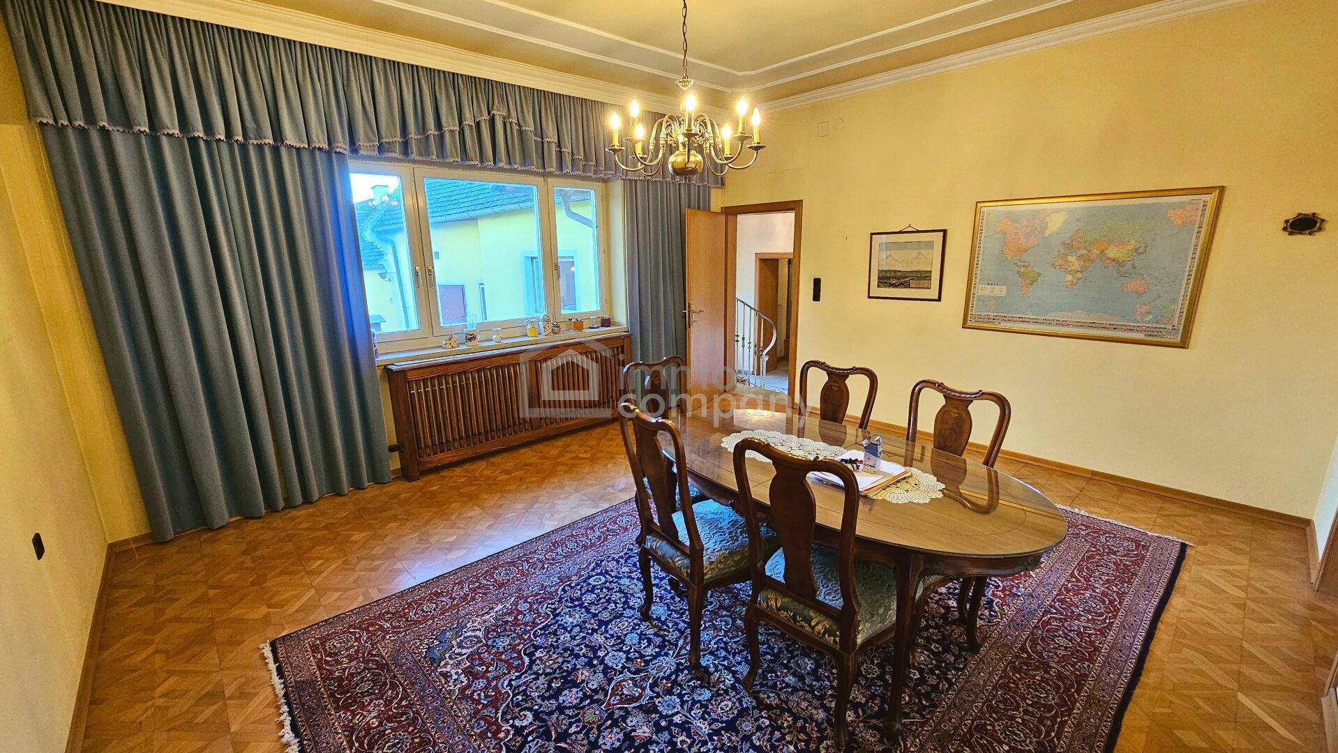 Klassisches Esszimmer mit ovalem Holztisch, Kronleuchter und großem Fenster mit blauen Vorhängen.