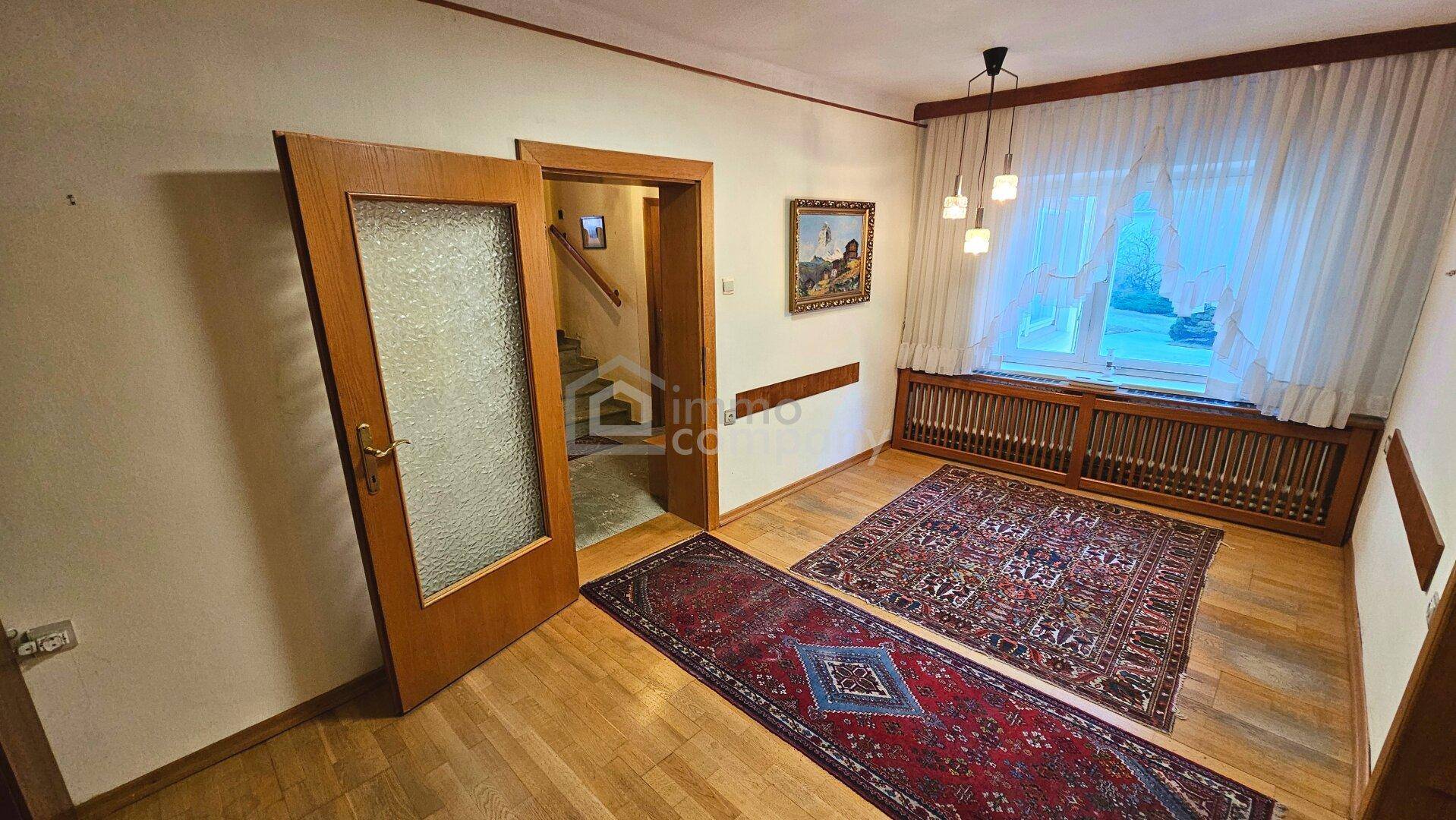 Helles Zimmer mit Holzfußboden, großem Fenster und Zugang zum Treppenhaus.