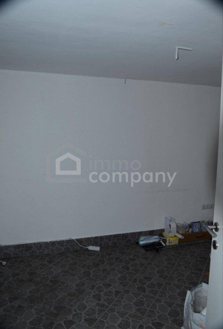 Leerer Raum mit weißen Wänden und Fliesenboden, einige Gegenstände liegen in einer Ecke.