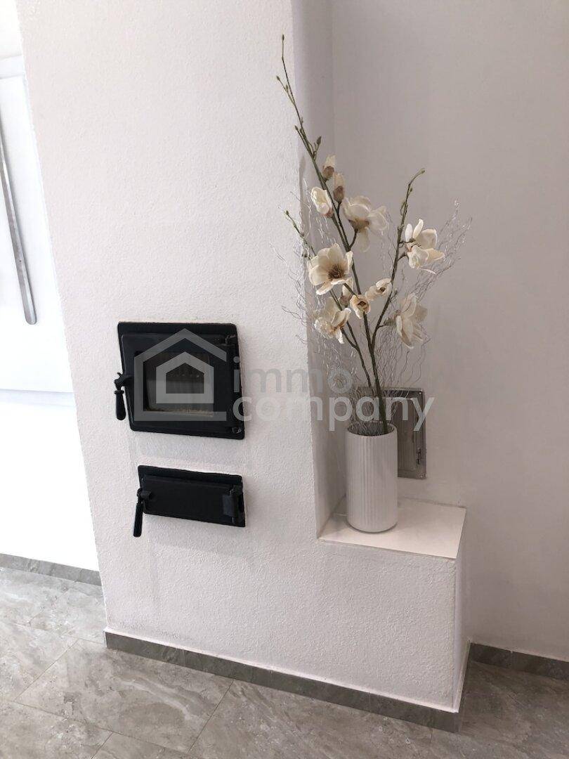 Weißer Kaminofen mit schwarzen Türen und einer dekorativen Vase mit Blumen.