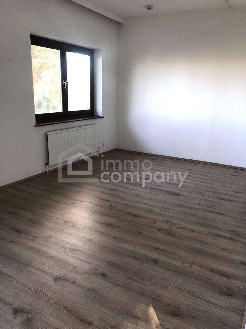 Leeres Zimmer mit hellem Laminatboden, weißen Wänden und einem Fenster mit Heizkörper darunter.