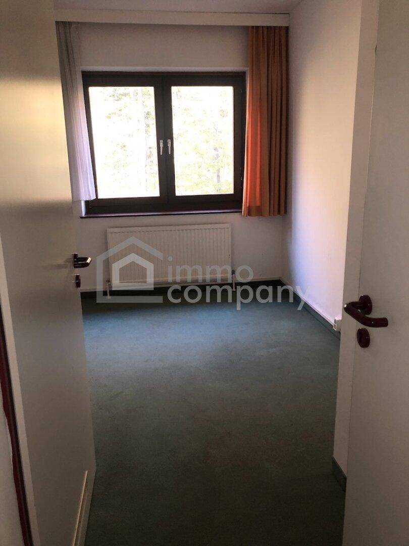 Leeres Zimmer mit grünem Teppichboden, weißen Wänden und einem Fenster mit Heizkörper.