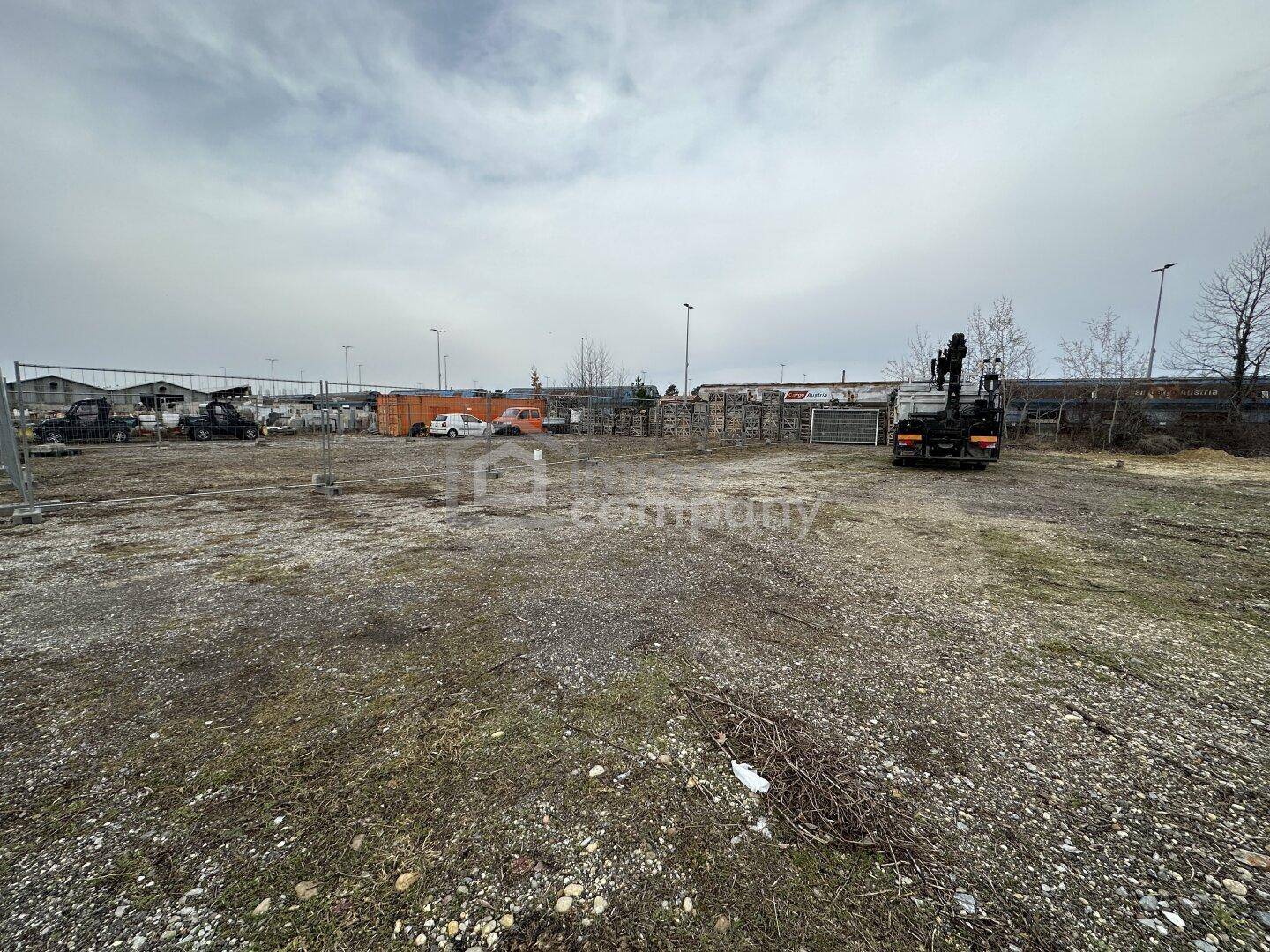 Weitläufiges, unbebautes Grundstück mit industriellen Elementen und Fahrzeugen im Hintergrund unter grauem Himmel.