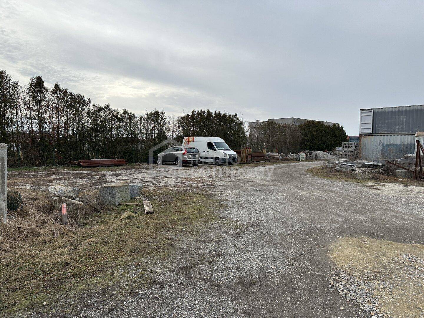 Schotterweg führt zu einem Bereich mit parkenden Fahrzeugen, umgeben von Bäumen und industriellen Strukturen.