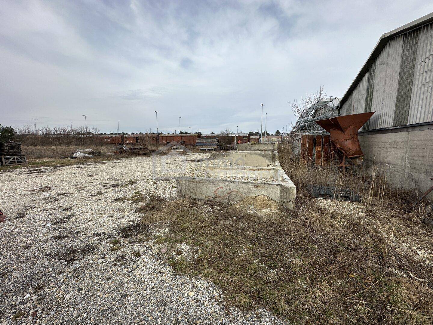 Außengelände mit Schotter, Betonstrukturen und einer Industriehalle im Hintergrund, umgeben von Unkraut und Ablagerungen.