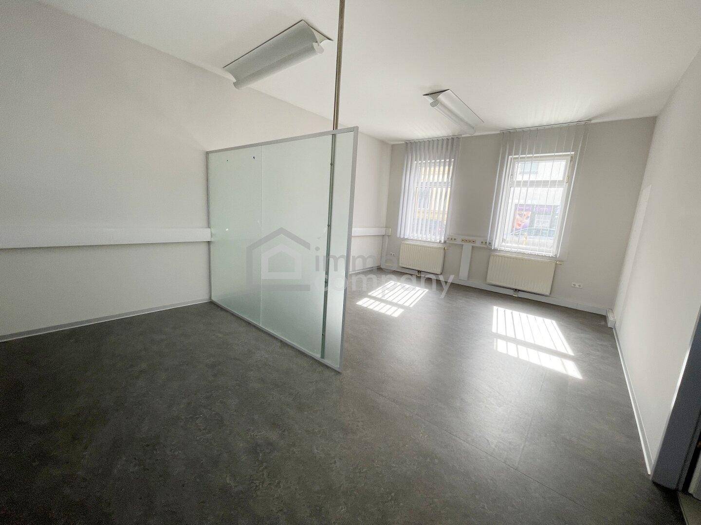 Leerer Büroraum mit dunklem Vinylboden, zwei Fenstern und einer modernen, halbhohen Trennwand.