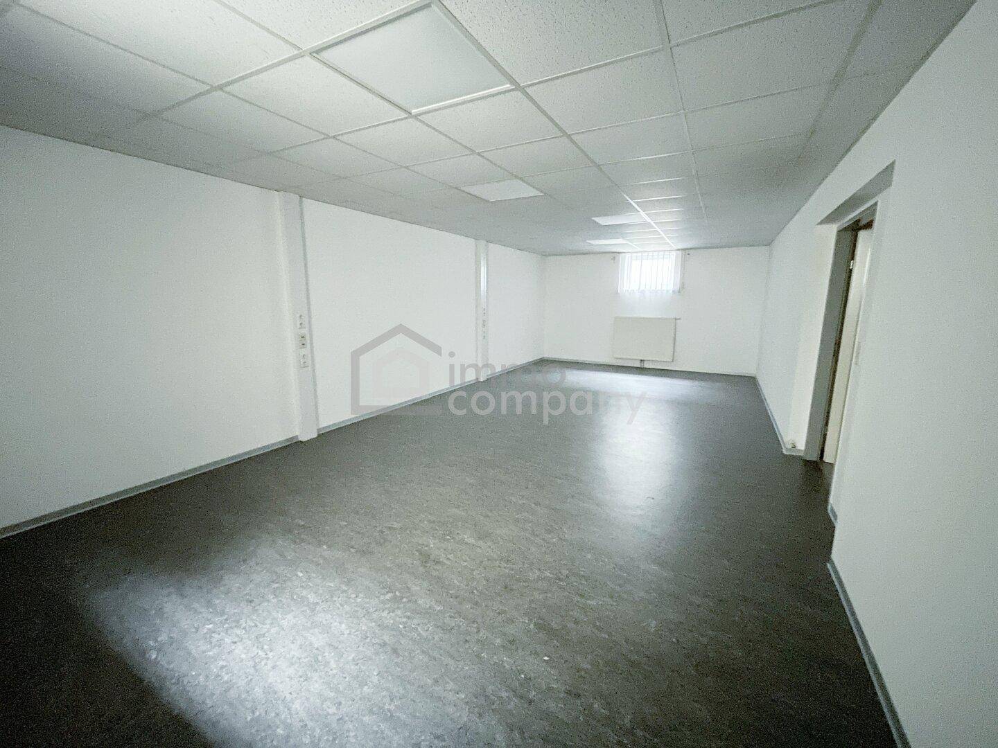 Großer, leerer Raum mit dunklem Vinylboden, abgehängter Decke und einem Fenster mit Heizkörper.