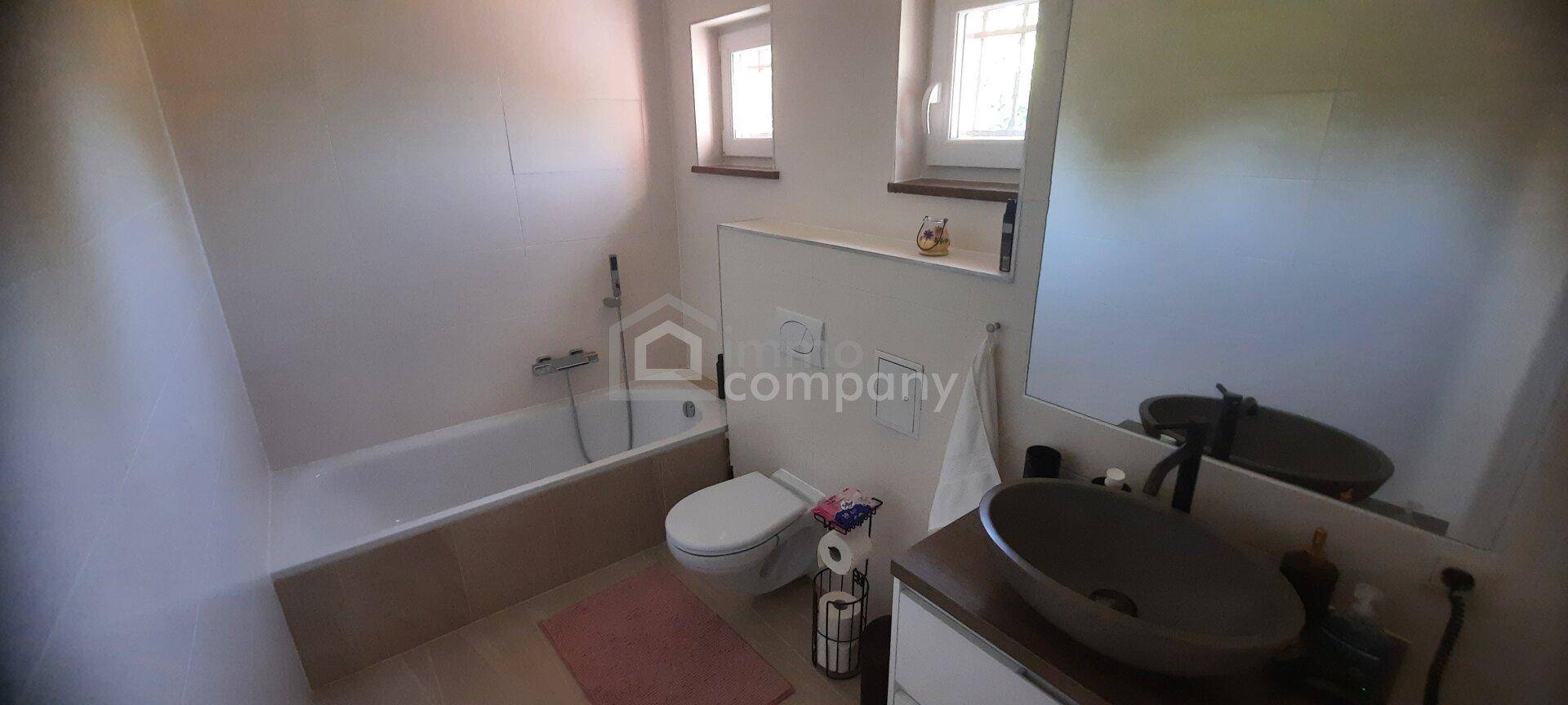 Helles Badezimmer mit Badewanne, WC und modernem Waschbecken unter zwei Fenstern.