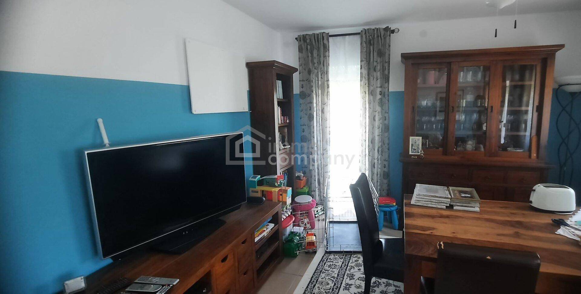 Wohnzimmer mit Fernseher, Holzmöbeln und einem großen Fenster, das viel Licht hereinlässt.