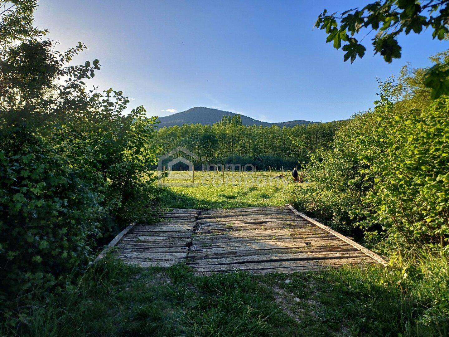 Rustikale Holzbrücke über einen Bach, umgeben von üppigem Grün und Bergen im Hintergrund.