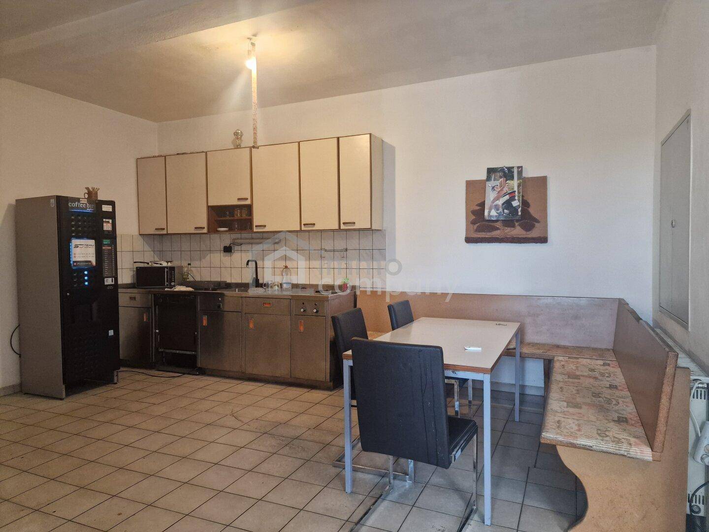 Einfache Küche oder Pausenraum mit Schränken, Spüle, Kaffeemaschine und Esstisch mit Sitzbank.