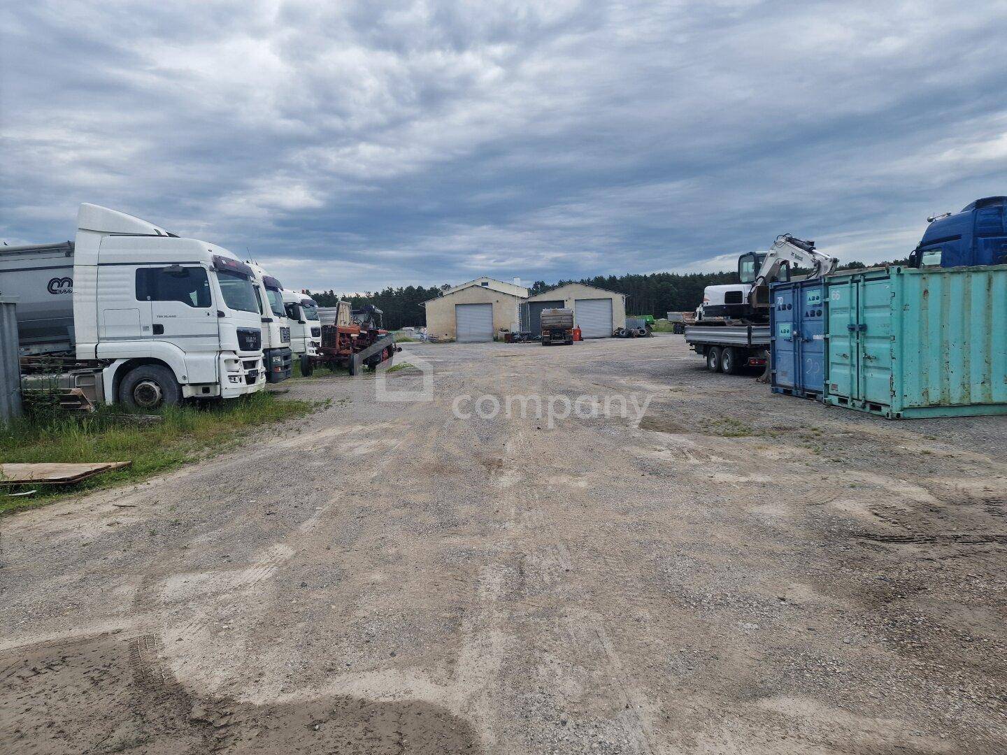 Großer Außenbereich mit Kiesfläche, Lastwagen, Containern und Lagerhallen im Hintergrund unter bewölktem Himmel.