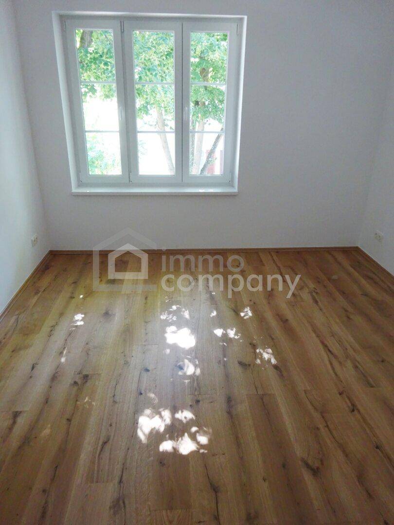 Heller Raum mit warmem Holzfußboden und einem großen Fenster, das viel natürliches Licht und einen grünen Ausblick bietet.