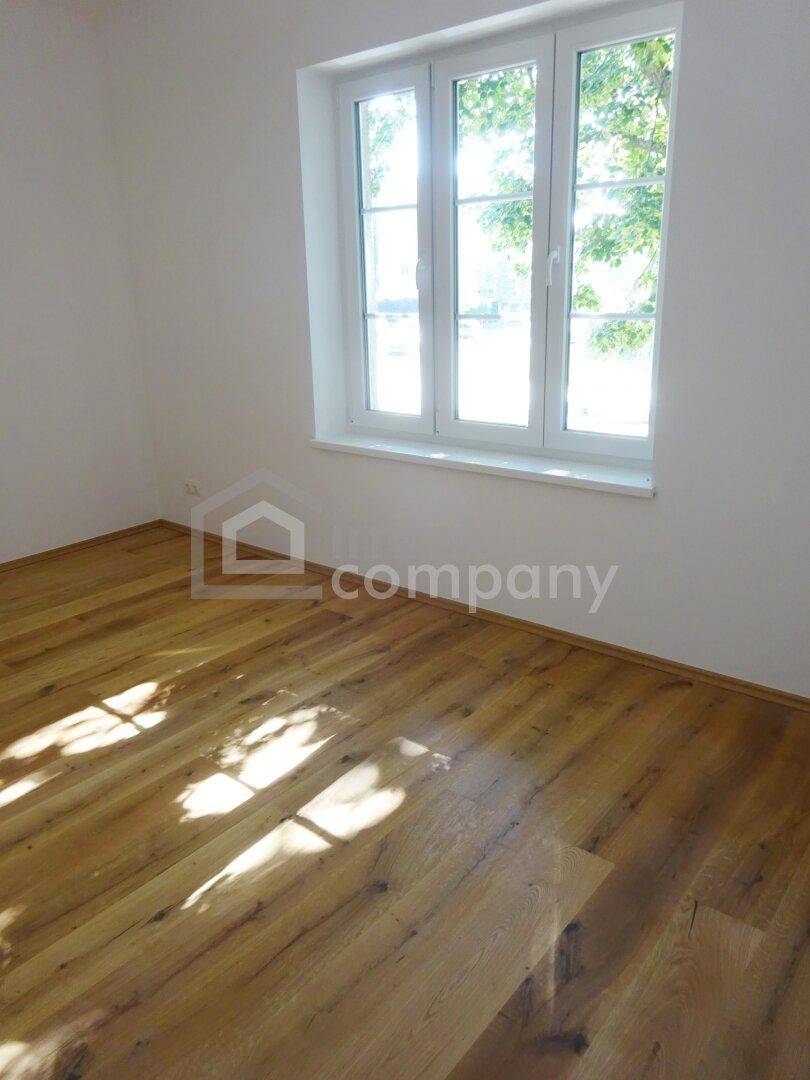 Lichtdurchfluteter Raum mit elegantem Holzboden und einem großen Fenster, das einen angenehmen Blick ins Grüne ermöglicht.