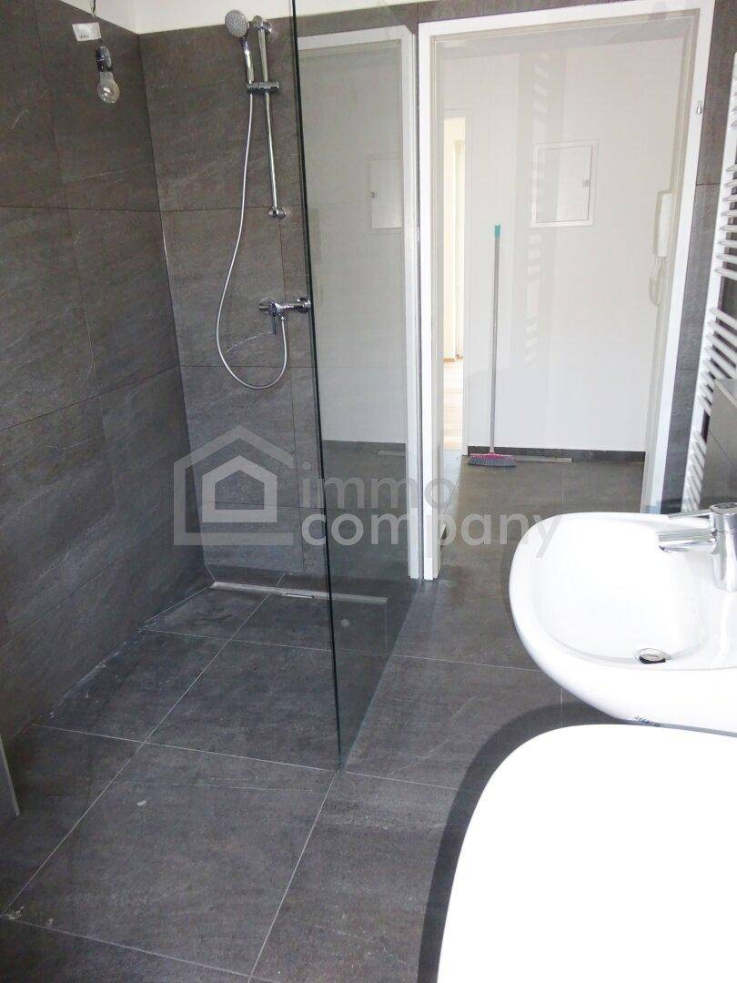 Geräumige, ebenerdige Dusche mit Glasabtrennung und modernen Fliesen im Badezimmer, daneben ein Waschbecken.