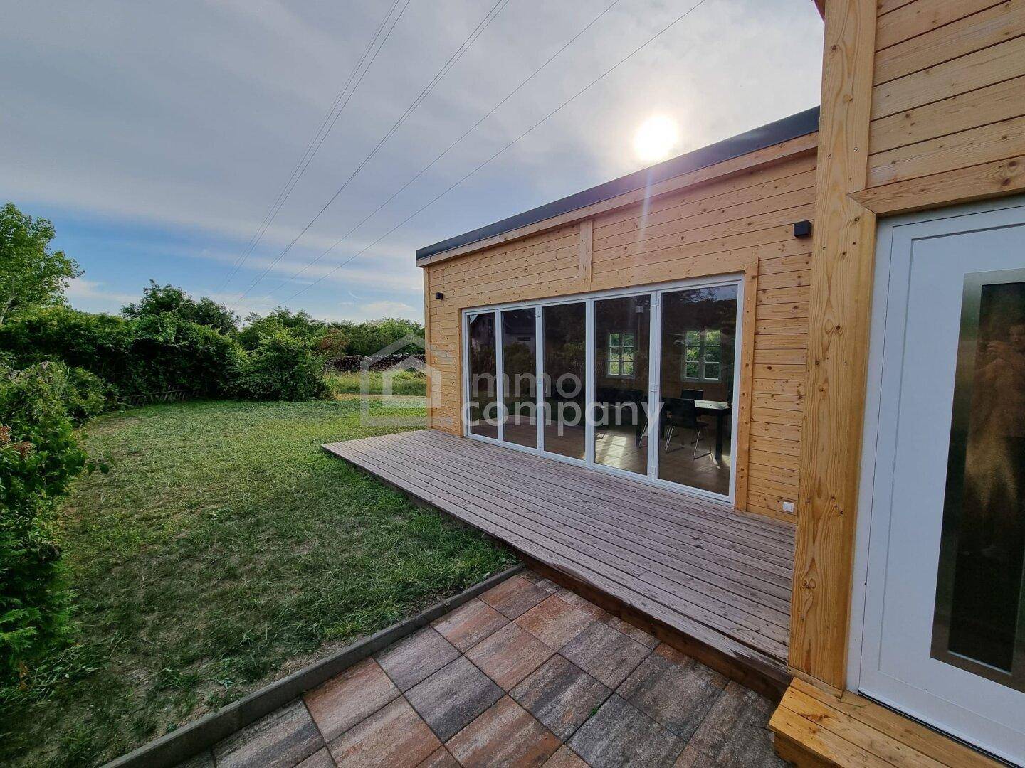 Holzhaus mit großer Holzterrasse und Garten, umgeben von viel Grün unter sonnigem Himmel.