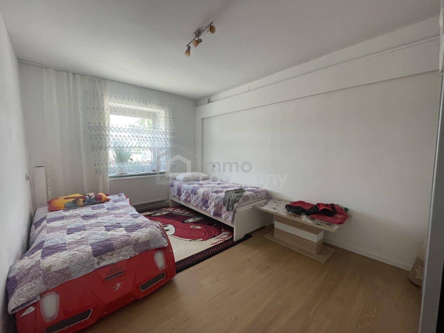 Kinderzimmer mit zwei Einzelbetten, hellem Holzfußboden und Fenster mit Vorhängen.