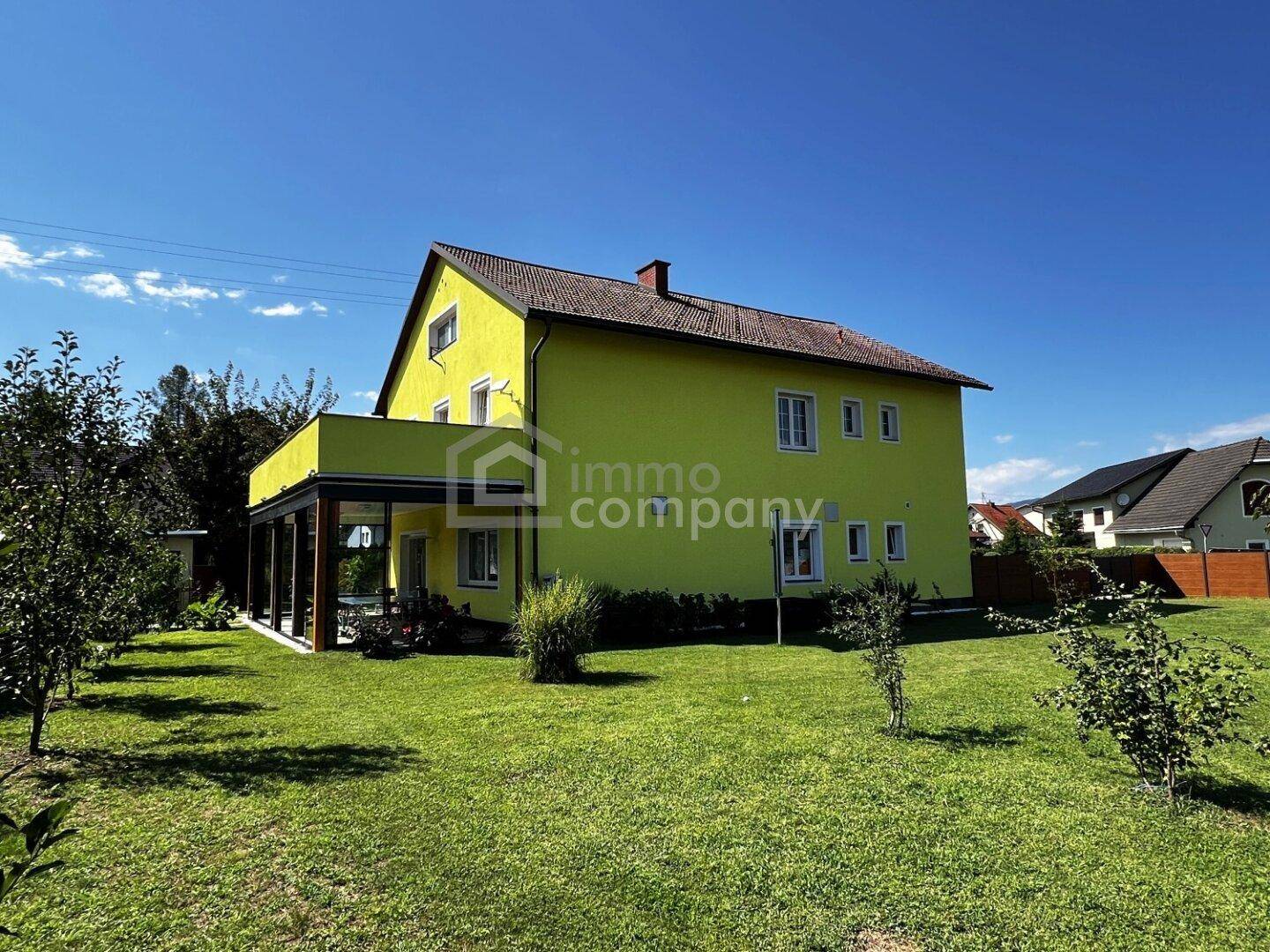 Rückansicht des gelben Hauses mit weitläufigem Rasen, jungen Bäumen und blauem Himmel.