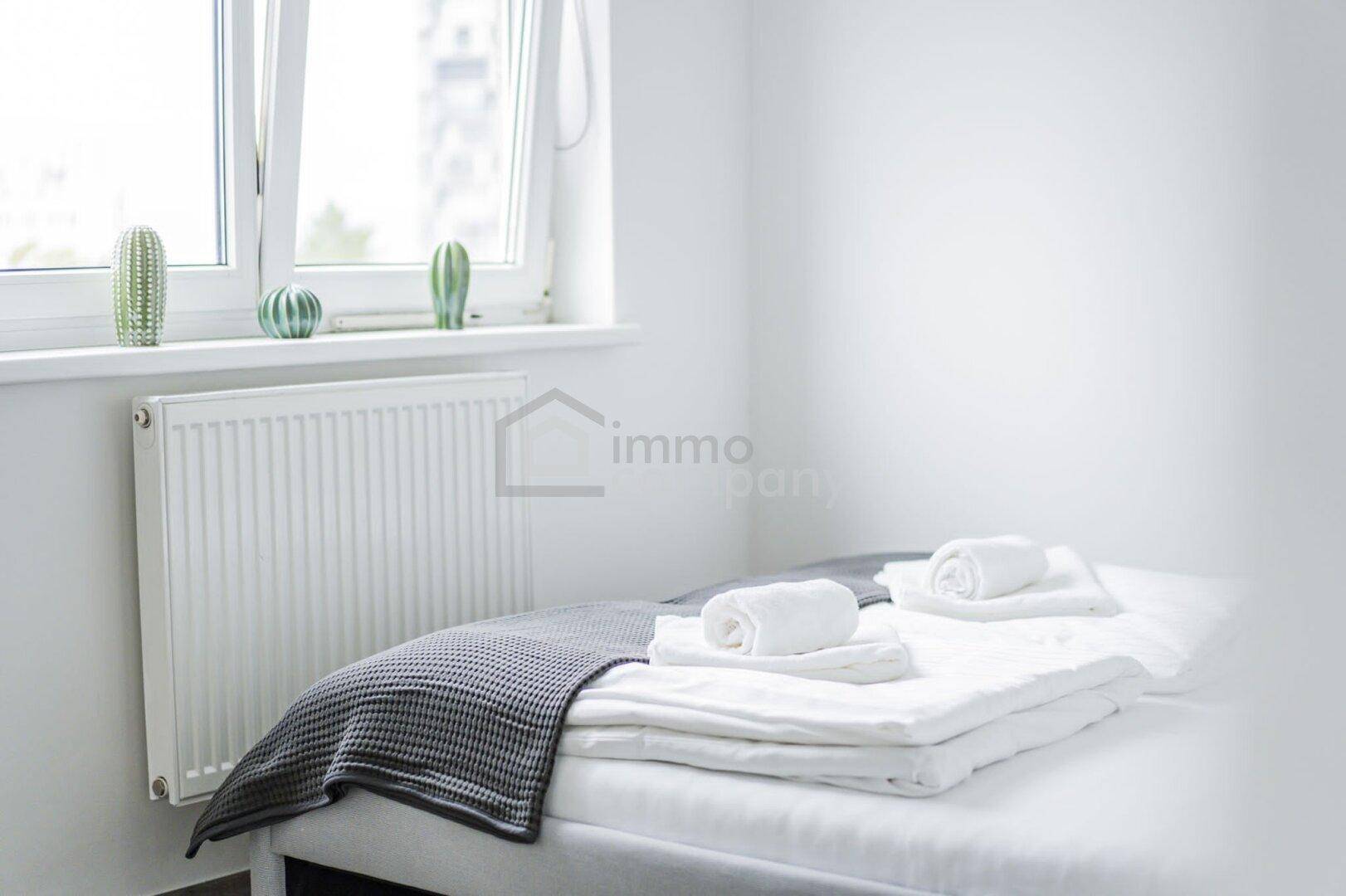 Schlichtes Schlafzimmer mit Einzelbett, Fenster und Heizkörper, in hellen Farbtönen gehalten.