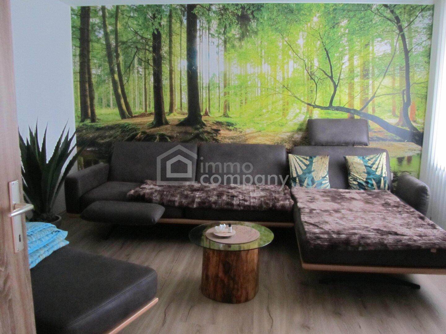 Gemütliches Wohnzimmer mit großem L-förmigem Sofa, Baumstamm-Couchtisch und Waldmotiv-Tapete.
