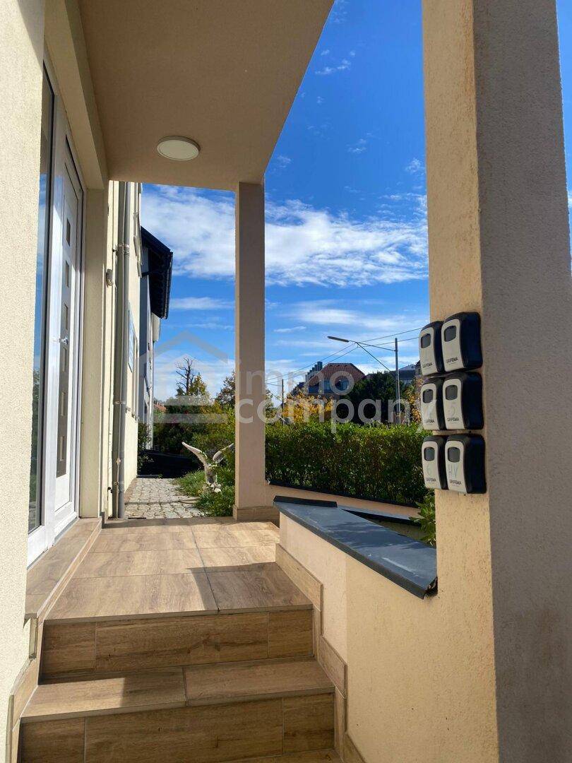 Überdachter Eingangsbereich mit Treppe und Säulen, umgeben von grünen Hecken.