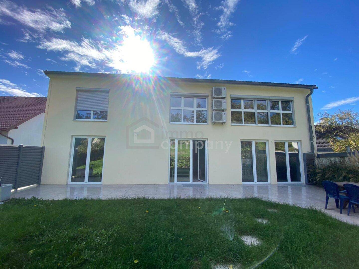 Rückseite des Hauses mit großer gepflasterter Terrasse, Rasenfläche und viel Sonnenlicht.