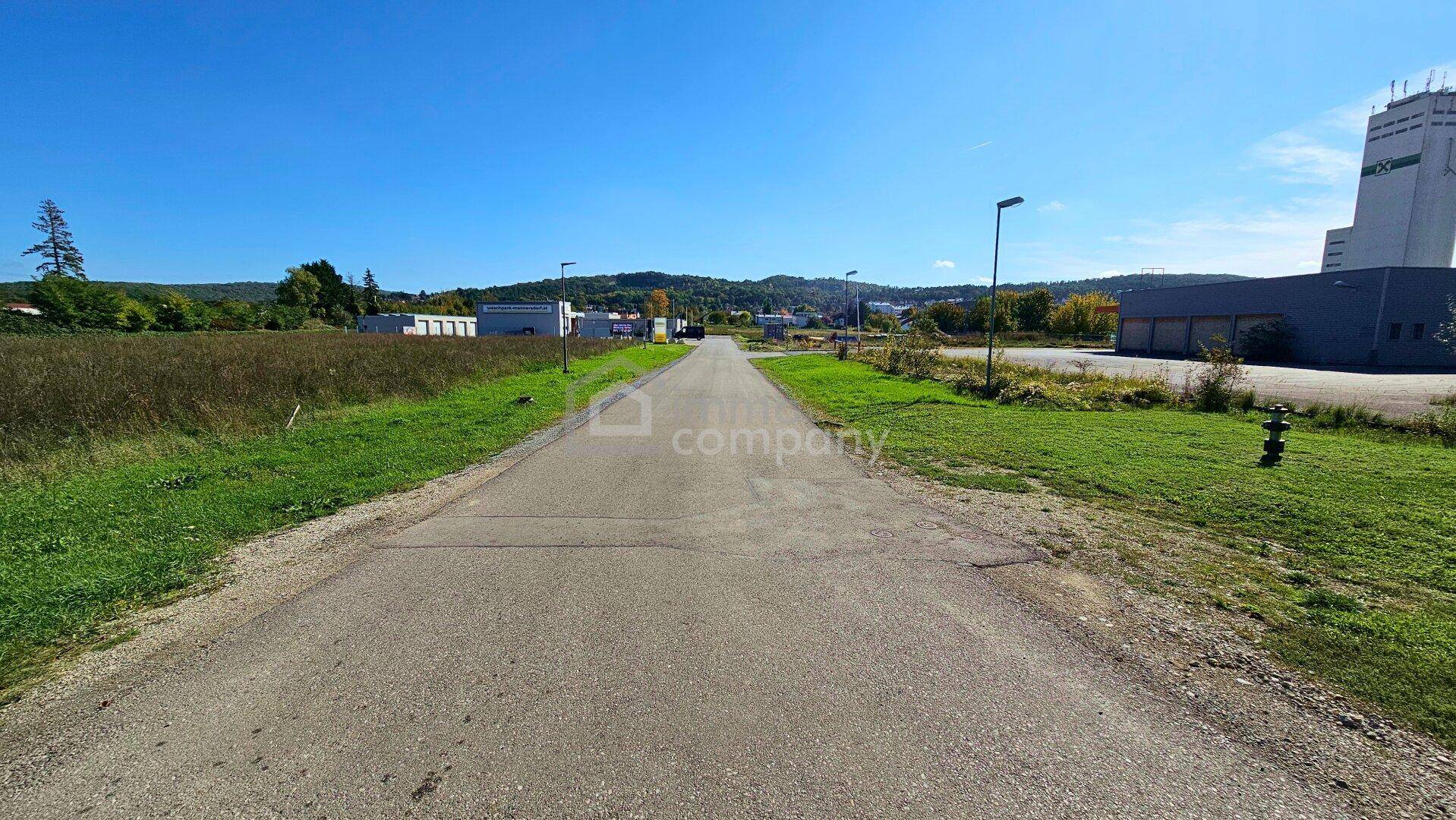 Eine asphaltierte Straße führt durch ein Gewerbegebiet mit Gebäuden und grünen Flächen unter blauem Himmel.