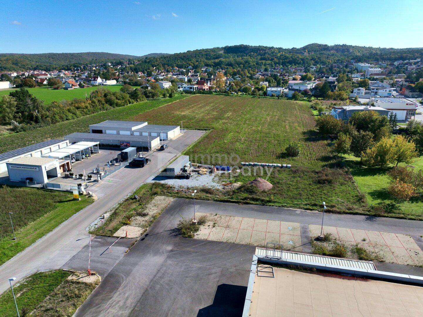 Luftaufnahme eines Gewerbegebiets mit bestehenden Gebäuden, Parkplätzen und einer großen unbebauten Fläche.