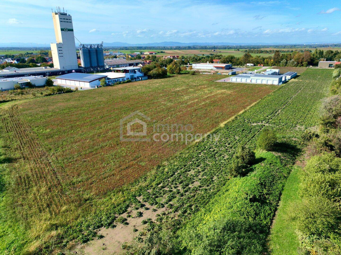 Weitläufiges Feld mit einigen Gebäuden im Hintergrund, umgeben von grüner Natur unter klarem Himmel.