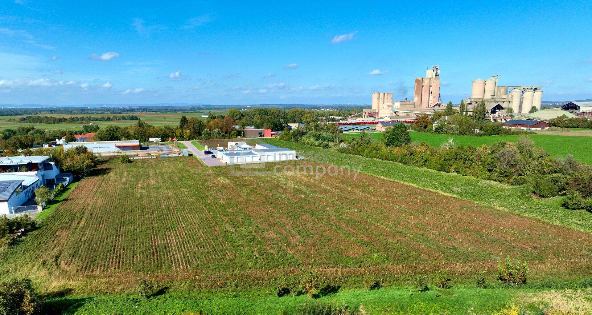 Weitläufiges Feld mit industriellen Gebäuden und Silos in der Ferne, umgeben von grüner Landschaft.