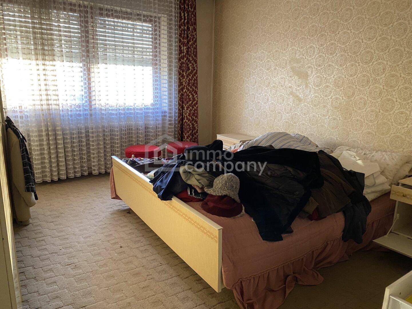 Schlafzimmer mit einem Bett, gemusterter Tapete und einem großen Fenster mit Jalousien.