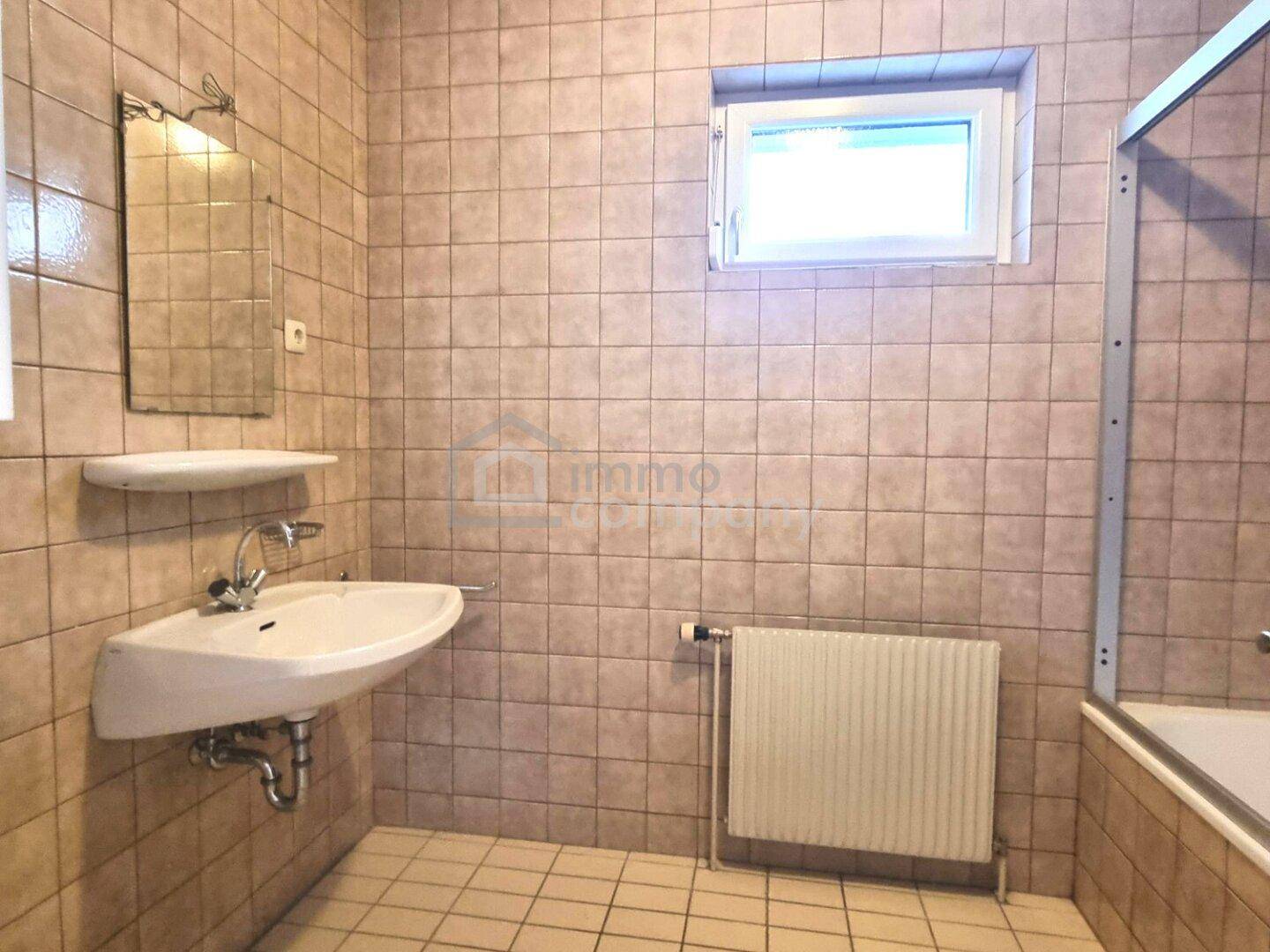 Gefliestes Badezimmer mit Waschbecken, Spiegel, Heizkörper und Badewanne mit praktischer Duschabtrennung.