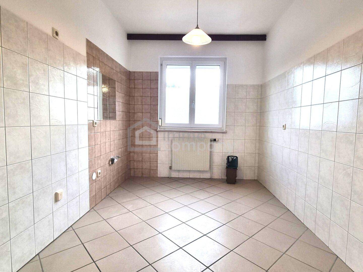 Leerer Raum mit Fliesenboden und gefliesten Wänden, Fenster und sichtbaren Anschlüssen für eine Küche.