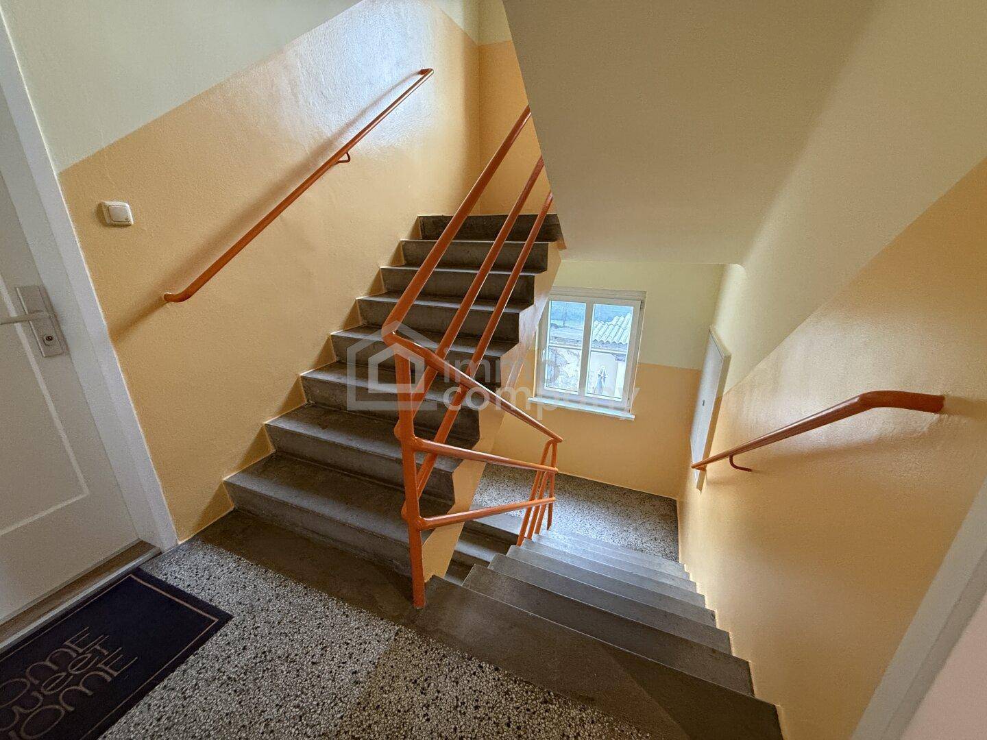 Treppenhaus mit orangefarbenem Geländer und Fenstern, das die verschiedenen Etagen verbindet.