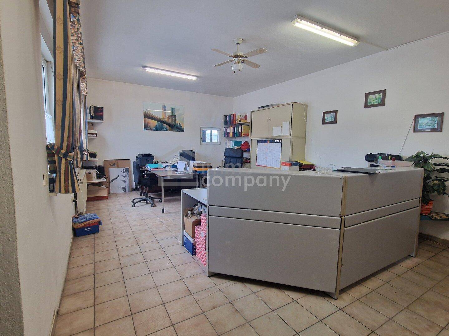Geräumiges Büro mit mehreren Arbeitsplätzen, Deckenventilator und Fliesenboden.