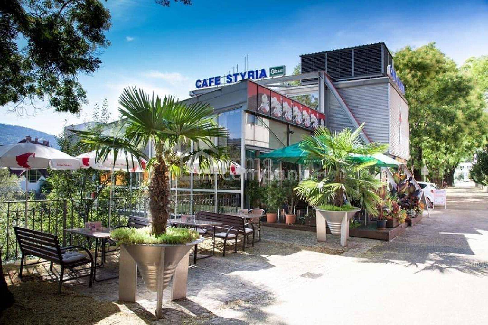 Attraktives Café-Gebäude mit Außenterrasse, Palmen und Sonnenschirmen in grüner Umgebung.