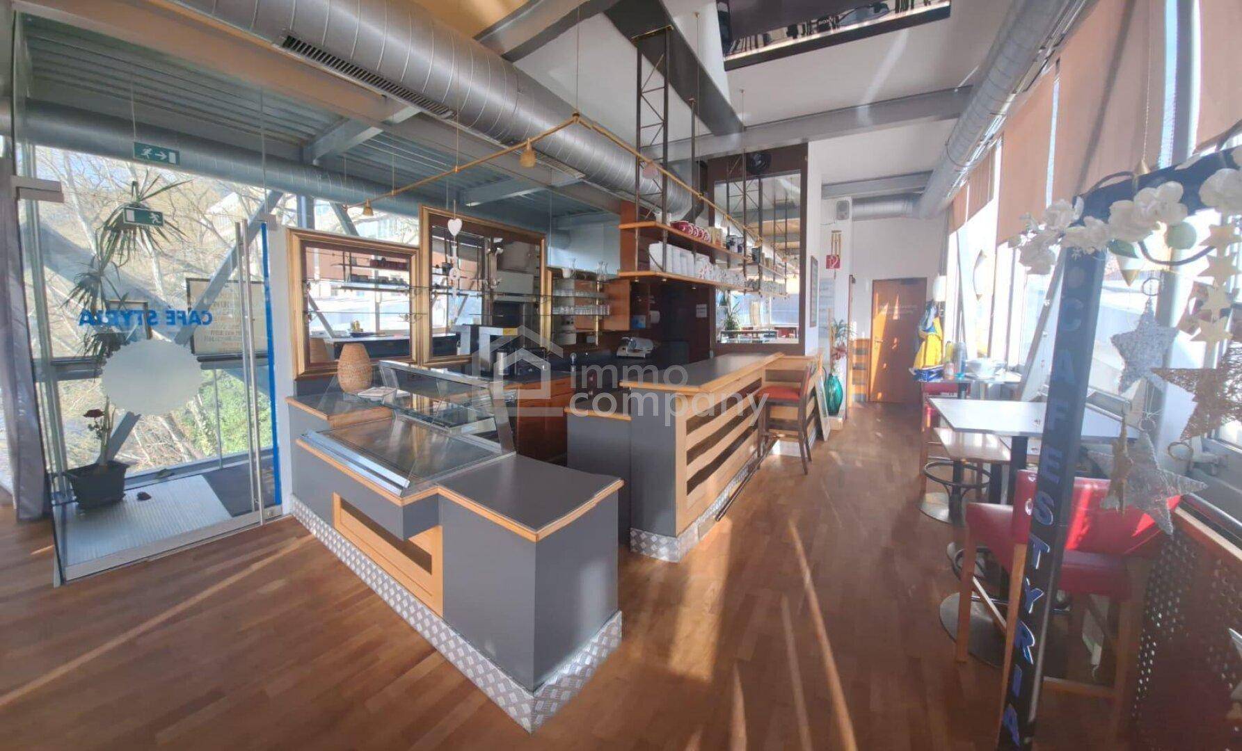 Offener Barbereich mit Theke, Kühlvitrine und Holzboden, umgeben von großen Glasflächen.