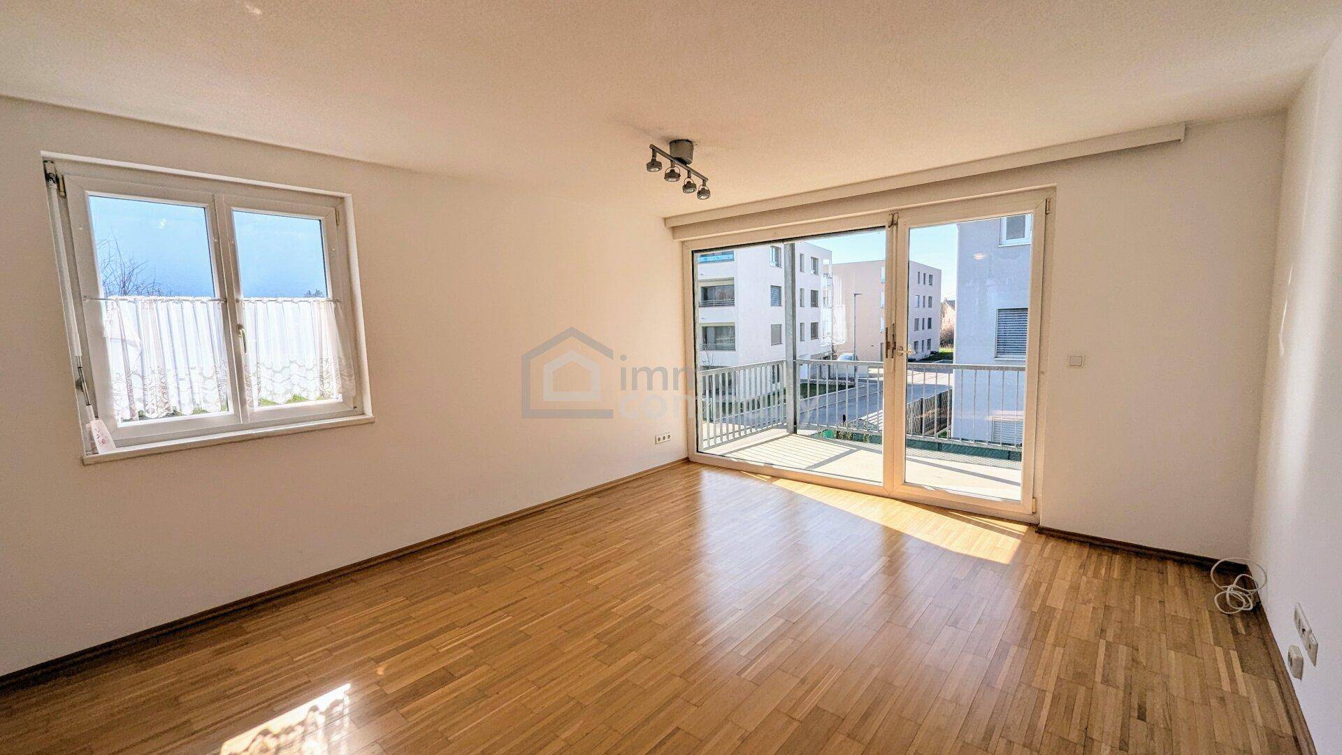 Geräumiges, leeres Wohnzimmer mit Holzboden und großen Fenstern, die viel Tageslicht hereinlassen und Zugang zum Balkon bieten.