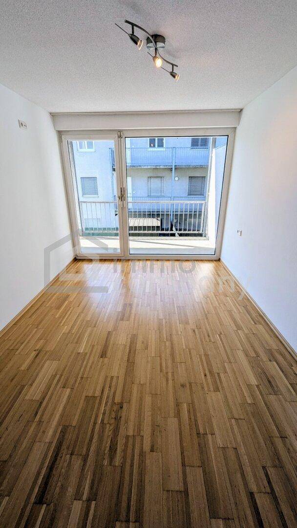 Leeres Schlafzimmer mit Holzboden und großen Fenstern, die Zugang zum Balkon bieten und den Raum hell erscheinen lassen.