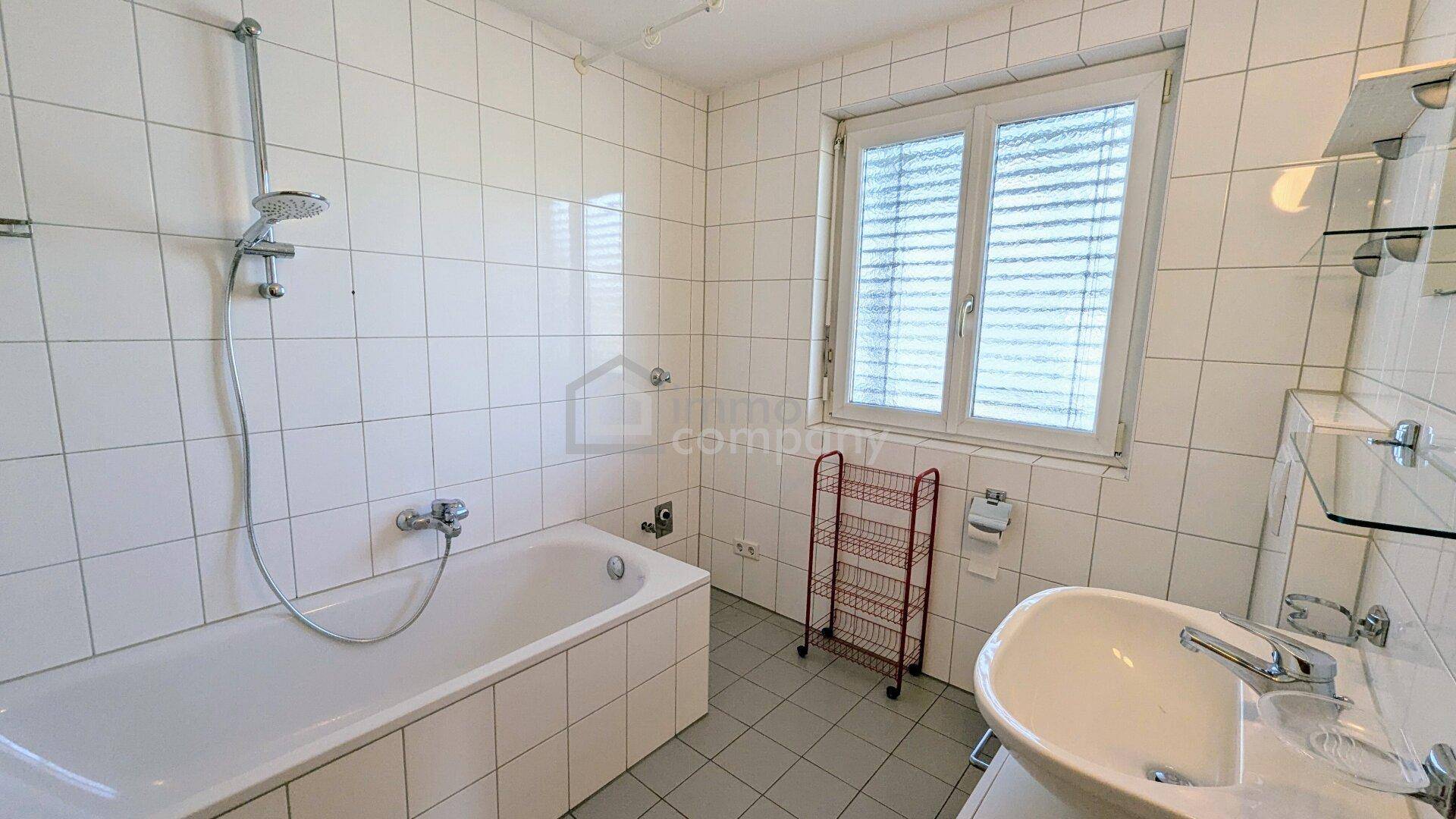 Helles Badezimmer mit weißen Fliesen, Badewanne mit Duschfunktion und einem Fenster für gute Belüftung.