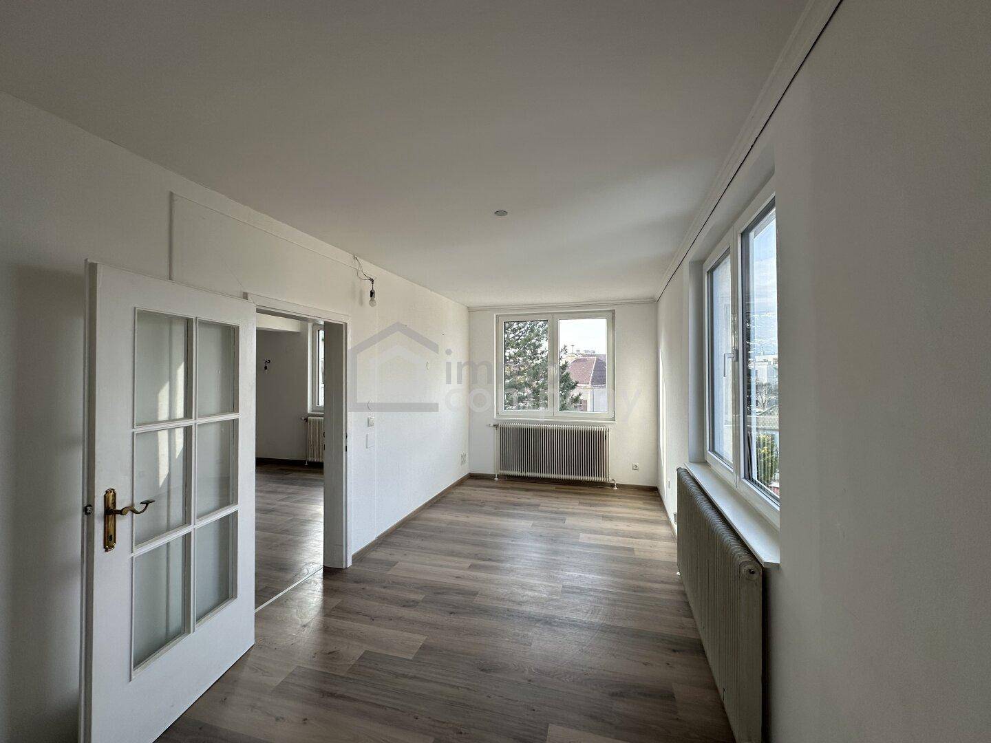 Helles Zimmer mit Laminatboden und großen Fenstern, die viel Tageslicht hereinlassen.