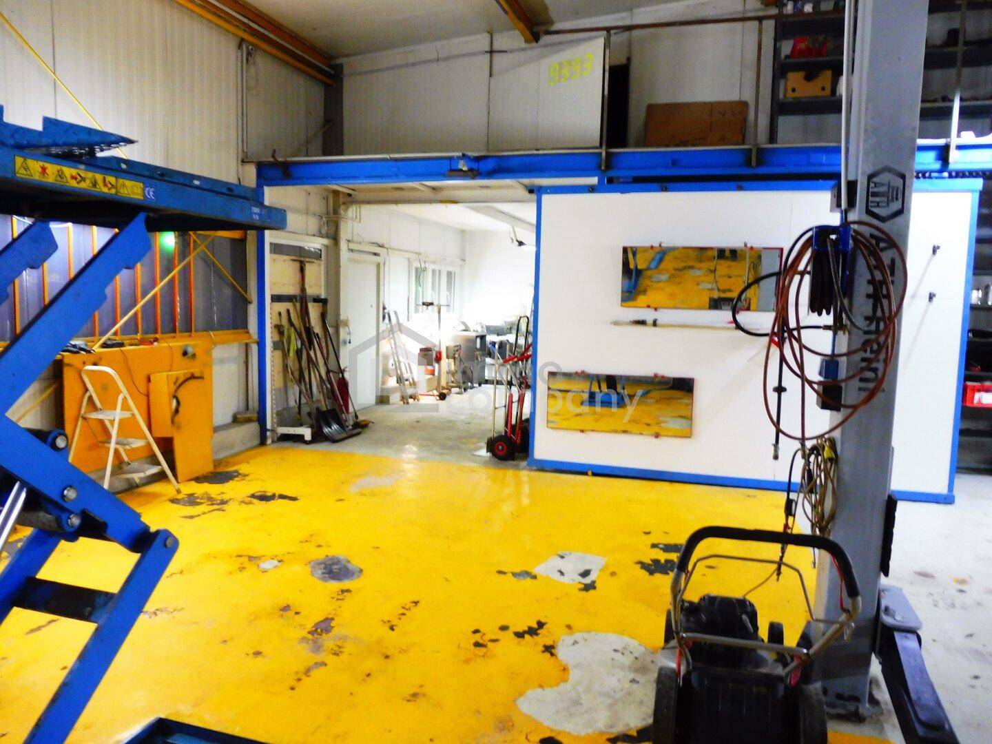 Werkstattbereich mit leuchtend gelbem Betonboden, einer Hebebühne und einem Durchgang zu einem weiteren Raum.